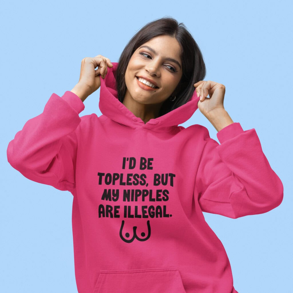 Woman wearing a heliconia pink hoodie sweatshirt with the text 'I'd be topless, but my nipples are illegal' printed on the front. There is a ling drawing of breasts under the text.