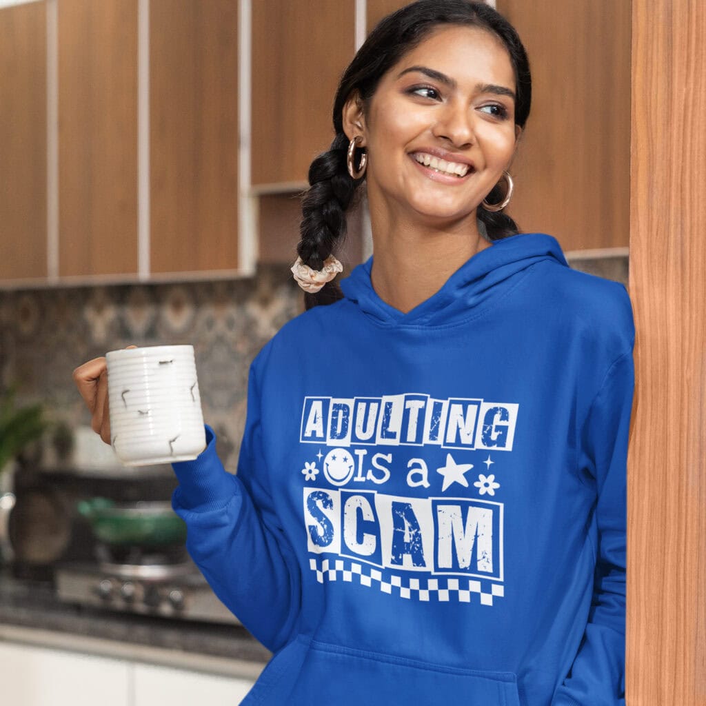 Woman wearing a royal blue hoodie sweatshirt with bold white text reading ‘Adulting is a Scam’ in mixed block and distressed fonts. The design includes a smiley face, star, and flower icons, with a checkerboard stripe along the bottom.