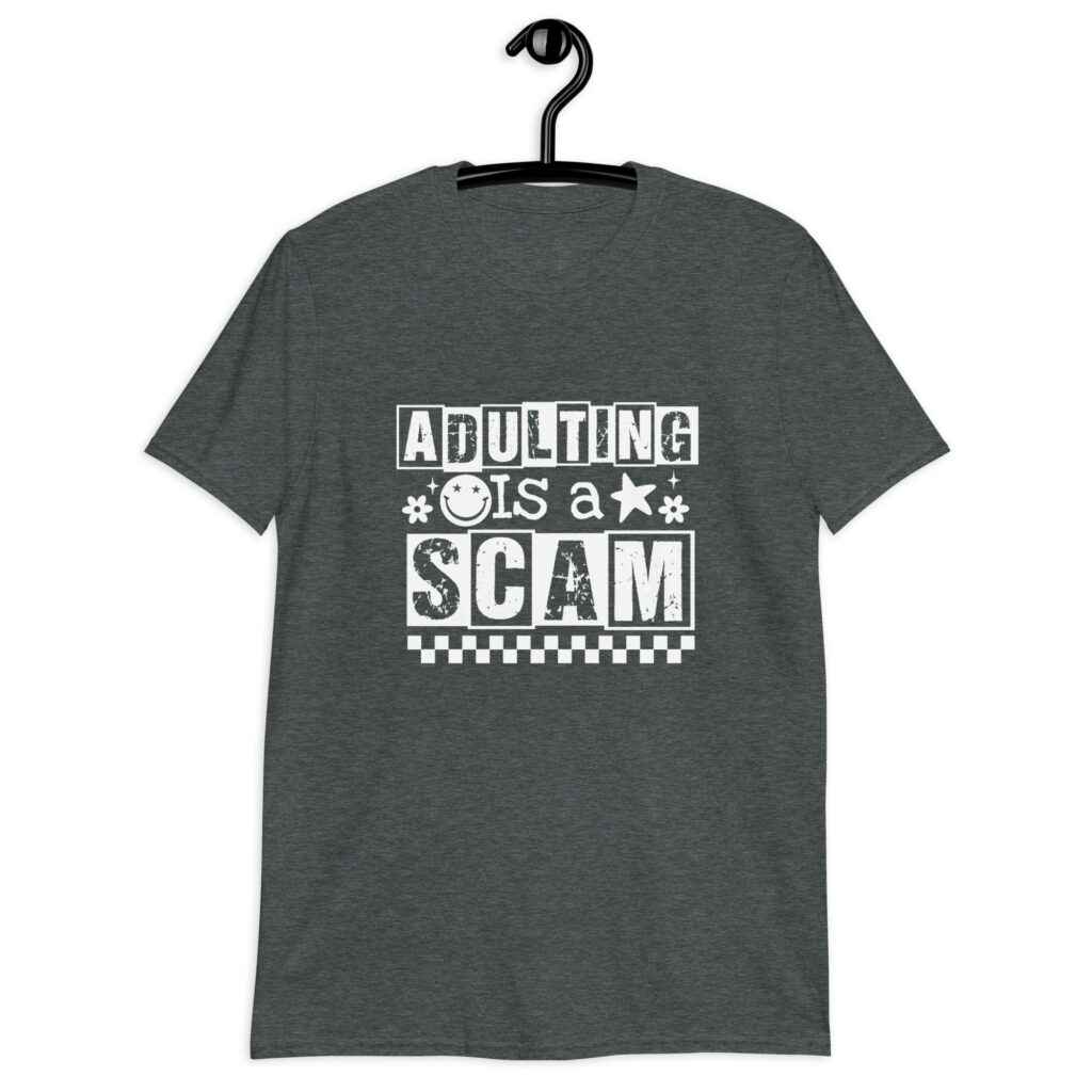 Dark heather grey t-shirt with bold white text reading ‘Adulting is a Scam’ in mixed block and distressed fonts. The design includes a smiley face, star, and flower icons, with a checkerboard stripe along the bottom.