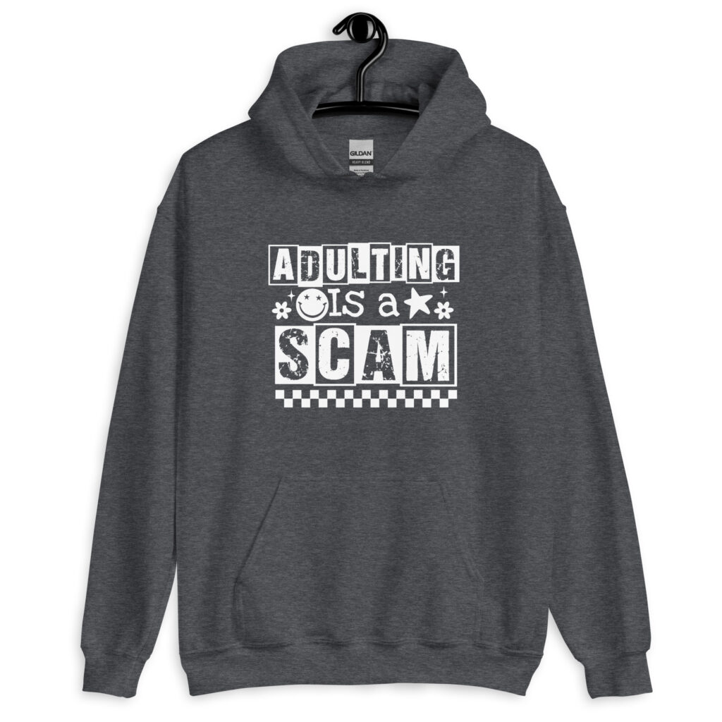 Dark heather grey hoodie sweatshirt with bold white text reading ‘Adulting is a Scam’ in mixed block and distressed fonts. The design includes a smiley face, star, and flower icons, with a checkerboard stripe along the bottom.