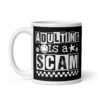White ceramic mug featuring a black wraparound design with bold distressed white text reading ‘Adulting is a Scam.’ The text includes a smiley face, star, and flower icons, with a white checkerboard stripe along the bottom.