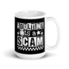 White ceramic mug featuring a black wraparound design with bold distressed white text reading ‘Adulting is a Scam.’ The text includes a smiley face, star, and flower icons, with a white checkerboard stripe along the bottom.
