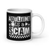 White ceramic mug featuring a black wraparound design with bold distressed white text reading ‘Adulting is a Scam.’ The text includes a smiley face, star, and flower icons, with a white checkerboard stripe along the bottom.